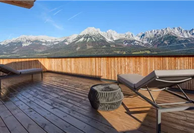 A terrace with two loungers and a table. In the background, majestic mountains with snow and a clear blue sky are visible.