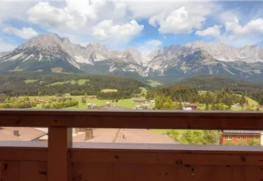 A breathtaking mountain landscape with green meadows and old houses in the foreground. The sky is clear with some clouds.