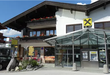 A modern building with a glass entrance area. In the foreground, various shops and a bank can be seen.