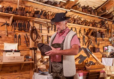 Ein Handwerker in traditioneller Kleidung betrachtet ein kunstvoll geschnitztes Holzstück. Im Hintergrund sind viele Werkzeuge an einer Holzwand sichtbar.