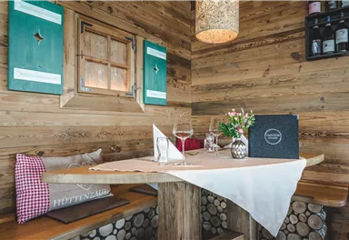 Ein gemütlicher Tisch in einer rustikalen Holzhütte mit einer blühenden Blumenmitte. Die Wände sind mit Holz verkleidet und grüne Fensterläden ergänzen die Atmosphäre.