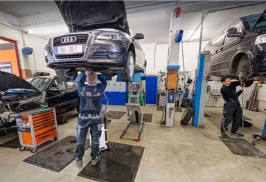 In einer Werkstatt werden zwei Autos von Hebebühnen gehalten. Mechaniker arbeiten an den Fahrzeugen und führen Reparaturen durch.