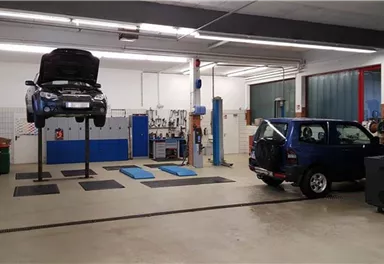 Eine Werkstatt mit zwei Autos, eines ist auf einer Hebebühne. Der Boden ist sauber und die Wände sind hell.