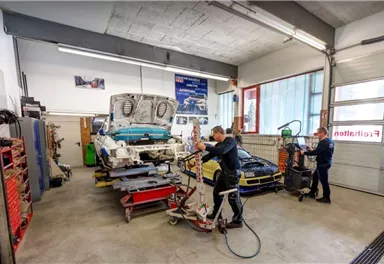 Eine Autowerkstatt mit zwei Mechanikern, die an Fahrzeugen arbeiten. Im Hintergrund sind Werkzeuge und Teile organisiert.