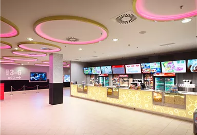 A modern cinema hall with a large selection of snacks and drinks. The walls are lit with pink lights and there are several screens with movie announcements.
