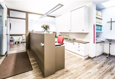 A modern reception area with wooden flooring and white furnishings. Floral decorations and a cross enhance the friendly atmosphere.