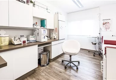 A modern treatment room with a red treatment table and bright furniture. The room has large windows that provide plenty of natural light.