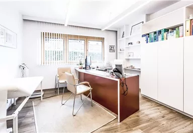 A modern office with a desk, a computer, and shelves. The walls are light-colored and there are windows with blinds.