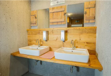Ein modernes Badezimmer mit zwei Waschbecken auf einer Holzablage. Im Hintergrund sind Spiegel und rustikale Holzkonturen zu sehen.