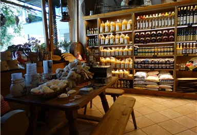 A cozy sales room with a wooden table and various regional foods on the table. In the background, there are shelves with jars full of jams, oils, and other products.
