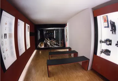 A modern exhibition space with exhibits on the walls and seating in the center. The lighting is dimmed, creating a cozy atmosphere.