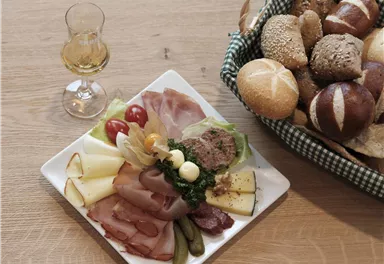 A plate with various types of sausage and cheese, pickled cucumbers, and cherry tomatoes. Next to it stands a glass and a basket with rolls.