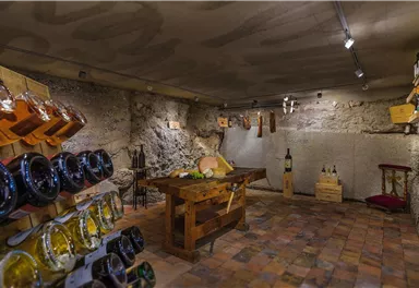 A cozy wine cellar with wooden shelves and bottles. In the center stands a wooden table, surrounded by stone walls.