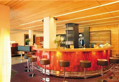 A modern bar with a round wooden counter and elegant bar stools. The decor is bright and friendly with warm wood and glass accents.