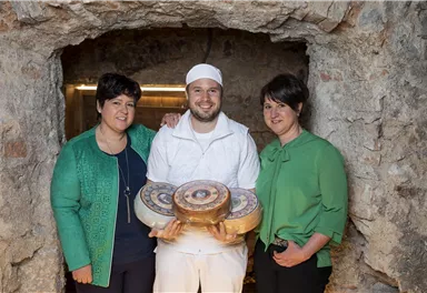 Eine Gruppe von drei Personen steht zusammen in einem Steinraum. Der Mann in der Mitte hält Käseräder, während die beiden Frauen lächeln.