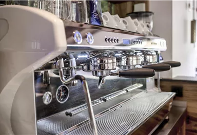 Eine moderne Espressomaschine mit glänzendem Metall und schwarzen Griffen. Im Hintergrund sind einige Tassen und das Interieur eines Cafés zu sehen.