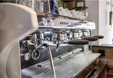 Eine moderne Espressomaschine mit glänzendem Metall und schwarzen Griffen. Im Hintergrund sind einige Tassen und das Interieur eines Cafés zu sehen.