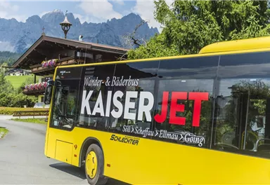 Ein gelber Bus mit der Aufschrift "KaiserJET" fährt durch eine malerische Landschaft. Im Hintergrund sind Berge und eine gemütliche Almhütte zu sehen.