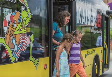 Eine Frau und zwei Mädchen steigen aus einem bunten Bus aus. Der Bus hat eine auffällige Gestaltung mit einer Zeichnung einer Froschfigur.