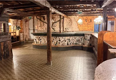 A rustic room with wooden walls and a tiled floor. In the center is a table and simple furnishings that create a cozy atmosphere.