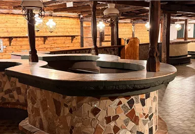 A rustic room with a large, round stone table in the center. The walls are made of wood and the floors are patterned.