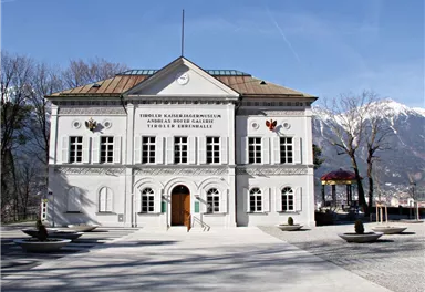 Ein historisches Gebäude mit klassischer Architektur und bunten Verzierungen. Im Hintergrund sind Bäume und ein klarer Himmel zu sehen.