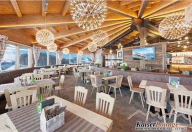A modern restaurant with wooden decor and large windows. The tables are set comfortably and the lighting is complemented by stylish pendant lights.