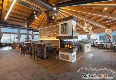 A cozy restaurant with rustic wood decor and large windows. The open fireplace in the center creates a warm atmosphere.