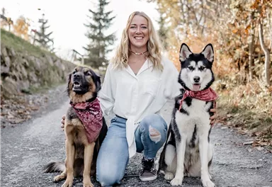 A woman is kneeling on a path and posing with two dogs. The dog on the left is brown and the dog on the right is black and white.