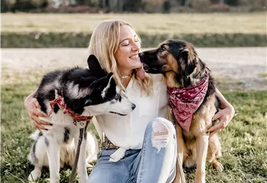 A woman is sitting on the grass and embracing two dogs. She smiles happily while the dogs are lovingly by her side.