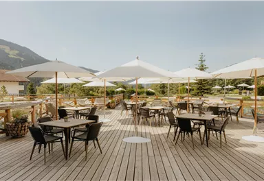 Eine gemütliche Terrasse mit Holztischen und Stühlen. Große Sonnenschirme bieten Schatten und umgeben von grünen Landschaften.