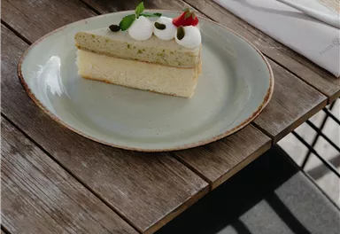 Ein Stück Kuchen mit einer cremigen Schicht und einem grünen Belag auf einem Teller. Im Hintergrund sind eine Tasse Kaffee und Besteck auf einem Holztisch zu sehen.
