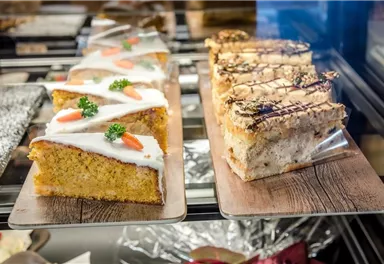 Eine Auswahl an köstlichen Kuchen in einer Vitrine. Links ein karottenförmiger Kuchen mit Glasur und Dekoration, rechts ein schnittiger Kuchen mit Schokolade und Nüssen.