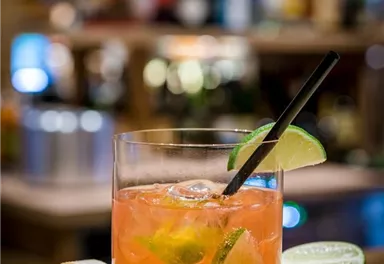 Ein erfrischender Cocktail in einem Glas, garniert mit Limettenscheiben. Im Hintergrund ist eine gemütliche Baratmosphäre zu sehen.