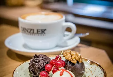 Eine Tasse Kaffee steht neben einem Teller mit drei leckeren Pralinen und roten Johannisbeeren. Das Ambiente ist gemütlich und einladend.