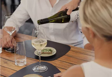 Ein Paar sitzt an einem Tisch und genießt ein Abendessen. Eine Person gießt Wein in ein Glas, während kleine Teller mit Speisen auf dem Tisch stehen.