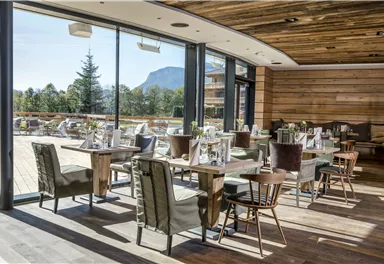 Ein modernes Restaurant mit großen Fenstern und Blick auf die Natur. Gemütliche Tische und Stühle sind in einer ansprechenden Umgebung angeordnet.