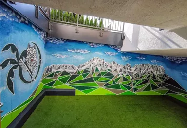 Eine bunte Wandmalerei mit einem Bergpanorama und blauen Himmel. Der Boden ist mit grünem Kunstrasen bedeckt.