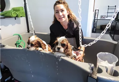 Eine Frau steht neben zwei Welpen, die in einer großen Wanne sitzen. Die Wanne wird mit Wasser gefüllt, während die Frau die Tiere betreut.