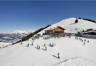 Eine schneebedeckte Berglandschaft mit vielen Skifahrern. Im Hintergrund befindet sich eine gemütliche Berghütte.