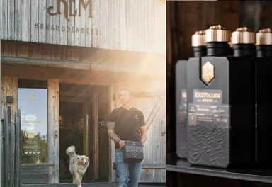A man is carrying a box of products in front of a wooden building. A dog is running beside him while the sun is shining.
