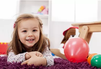 Ein fröhliches Mädchen liegt auf einem lila Teppich und lächelt. Bunte Spielzeuge und ein roter Ball sind um sie herum.