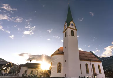 Eine malerische Kirche mit einem spitzen Turm und einer leuchtenden Sonne im Hintergrund. Der Himmel ist klar und hat eine schöne Abendstimmung.