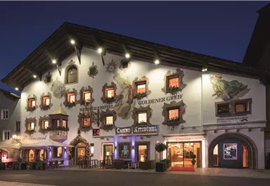 A traditional building with an ornate façade and colorful windows at dusk. In front of the building are illuminated entrances and an inviting atmosphere.