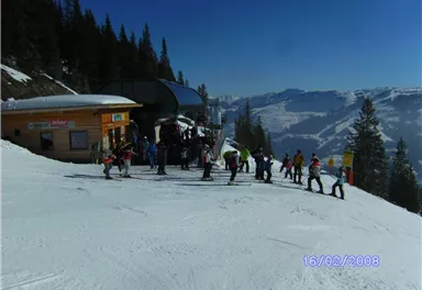 Eine belebte Skipiste mit Menschen, die auf einen Skilift warten. Im Hintergrund sind schneebedeckte Berge und ein klarer blauer Himmel zu sehen.