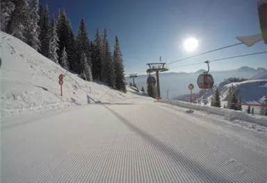 Eine verschneite Skipiste mit perfekt präpariertem Schnee. Im Hintergrund sind Skilifte und ein klarer blauer Himmel zu sehen.