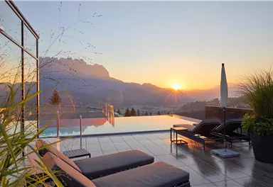 Ein wunderschöner Poolbereich mit Liegen und Blick auf die Berge. Die Sonne geht hinter den Bergen unter und taucht die Szene in warmes Licht.
