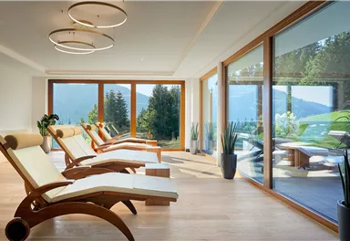 Ein entspannender Wellnessbereich mit gemütlichen Liegen und großen Fenstern. Durch die Fenster ist die Aussicht auf die grüne Landschaft und die Berge sichtbar.