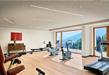 Ein modernes Fitnessstudio mit verschiedenen Trainingsgeräten. Große Fenster bieten einen Ausblick auf die Berge.