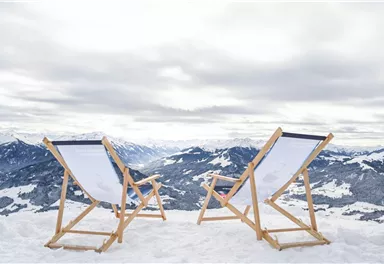 Zwei Liegestühle stehen auf schneebedecktem Boden mit Blick auf hochalpine Berge. Der Himmel ist bewölkt und die Landschaft ist winterlich und ruhig.